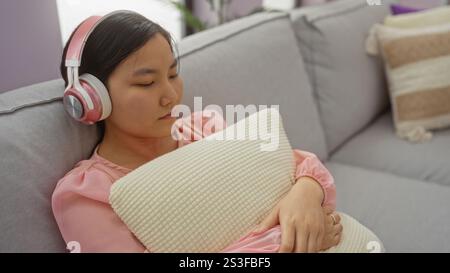 Frau, die sich mit Kopfhörern auf einer Couch in einem gemütlichen Wohnzimmer entspannt, rosa trägt, ein Kissen hält und eine friedliche Innenatmosphäre zu Hause genießt. Stockfoto