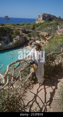 Junge hispanische Frau in weißer Kleidung und Sonnenhut genießt die Aussicht auf cala del moro, mallorca, spanien, umgeben von türkisfarbenem Wasser und felsigen Klippen. Stockfoto