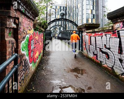 Manchester, Großbritannien, 01-04-2025: Person in Orange, die durch eine graffiti-bedeckte Stadtbrücke läuft. Stockfoto