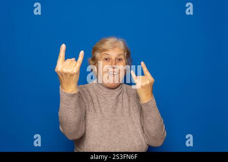 Eine weiße Frau, etwa 70 Jahre alt, trägt einen dunkelbeigen Pullover, der ein Symbol für ein Rock-on-Schild macht, mit Händen hoch, die breit lächeln, isoliert über Blau Stockfoto