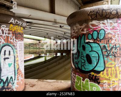 Salford , Manchester, Großbritannien, 01-04-2025: Unterführung mit lebendiger Graffiti-Kunst auf Betonsäulen durch einen reflektierenden Wasserweg. Stockfoto