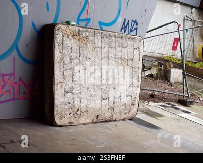 Salford , Manchester, Großbritannien, 01-04-2025: Eine abgenutzte Matratze lehnt sich an eine mit Graffiti bedeckte Stadtmauer unter einer Brücke. Stockfoto