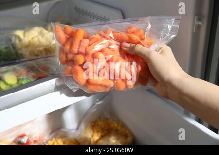 Frau nimmt Plastiktüte mit gefrorenen Karotten aus dem Kühlschrank, Großaufnahme Stockfoto