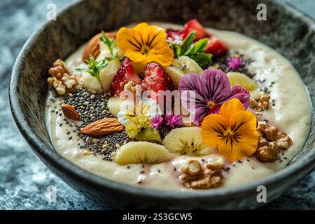 Eine lebendige Smoothie-Schüssel enthält geschnittene Bananen, Erdbeeren, Nüsse, Chiasamen und essbare Blumen, schön in einer Keramikschale angeordnet. Stockfoto