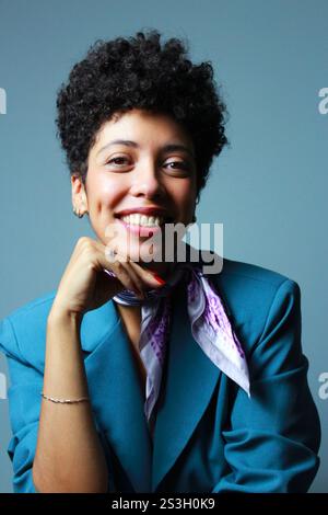 Erfolgreiche Geschäftsfrau, die hell in die Kamera lächelt, mit dem Kinn auf der Hand. Das Afro-weibliche Modell mit kurzen lockigen Haaren trägt einen blauen Blazer und einen lilafarbenen Schal. Stockfoto