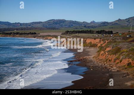 Wellen krachen sanft an einer sandigen Küste, umgeben von markanten Klippen und üppigem Grün mit Autos, die DEN HIGHWAY 1 im Hintergrund fahren. Die beschauliche Szene ca. Stockfoto