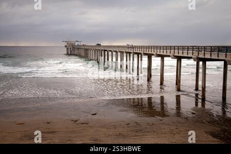 Der Pier in La Jolla erstreckt sich über den Ozean, wobei sanfte Wellen gegen die Stützpfeiler brechen. Der bewölkte Winterhimmel schafft eine ruhige Atmosphäre Stockfoto