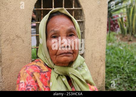 Eine traurig aussehende ältere Minangkabau-Frau, die einen grünen Hijab und ein Blumenkleid vor einer gelben Wand in Bukittinggi, West Sumatra, Indonesien trägt. Stockfoto