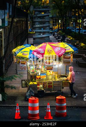 Ein lebendiger Hot Dog-Wagen in New York leuchtet in der Abenddämmerung in Manhattan. Diese Szene wurde vom MoMA-Fenster aufgenommen und zeigt die lebendige Street Food-Kultur der Stadt Stockfoto