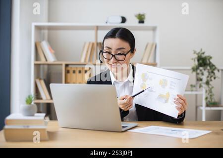 Selbstbewusste asiatische Geschäftsfrau mit Headset, die Daten während eines Videoanrufs über ein Notebook präsentiert. Frau mittleren Alters, die im Büro fernarbeitet und berufliche Fähigkeiten und Kommunikation unter Beweis stellt. Stockfoto