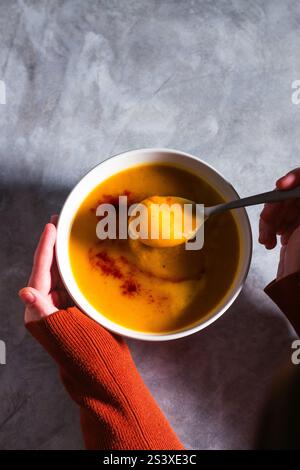 Frau, die Kürbissuppe mit Paprika-Gewürzen und Creme auf weißem Tisch probiert. Cremesuppe flach gelegt. Konzept der leckeren gesunden Ernährung. Stockfoto