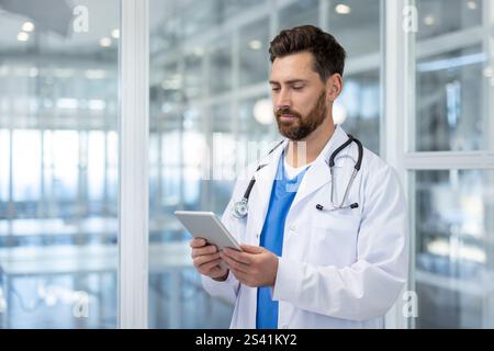 Ernsthafter erfahrener Arzt, der mit einem Tablet-Computer in der Hand in der Klinik steht. Reifer Mann im weißen medizinischen Mantel und mit einem Stethoskop unter Anwendung einer Applikation. Stockfoto