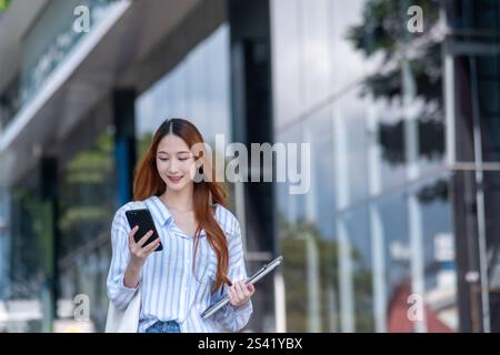 Die Frau läuft die Straße runter mit einem Handy in der Hand. Sie hält auch einen Laptop in der Hand Stockfoto