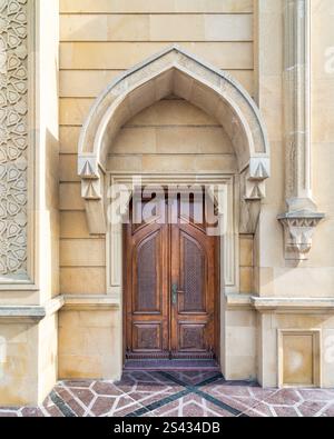 Eine wunderschön gefertigte Holztür, die von einem kunstvollen Bogengang eingerahmt wird, steht vor einer warmen Steinmauer. Sonnenlicht beleuchtet die Details und zeigt den einzigartigen architektonischen Stil Stockfoto