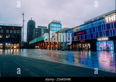 Stadtplatz in Enschede mit modernen Gebäuden, Geschäften und lebhaften Neonlichtern, die sich nach Regen auf nassem Gehweg spiegeln. Lebhafte urbane Atmosphäre in der Abenddämmerung Stockfoto