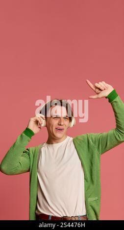 Mann, der Musik mit Kopfhörern genießt, mit der Hand spielerisch Gesten spielt, grüne Strickjacke und weißes T-Shirt vor weichem rosa Studiohintergrund trägt. Stockfoto