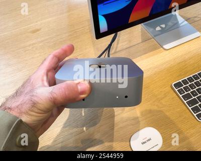 Paris, Frankreich - 10. Dezember 2024: Nahaufnahme einer Hand, die einen Apple Mac Mini in einem Geschäft hält, mit Monitor und Tastatur Stockfoto