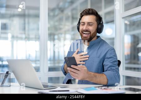 Mann mit Kopfhörern, der sich lustige und unterhaltsame Videos online auf dem Smartphone ansieht, Büroangestellte lacht, während er am Schreibtisch mit Laptop im Büro sitzt. Stockfoto