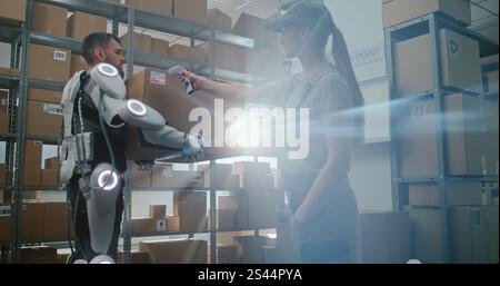 High-Tech Advanced Sorting Center: Lagerarbeiter im futuristischen Ganzkörper-Roboter-Exoskelett geht mit schwerer Kartonbox. Logistikspezialistin Scannt Paket Und Überprüft Versandinformationen. Stockfoto