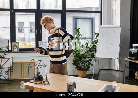 Ein junger Mann in einem gestreiften Pullover überprüft sein Handy in einem schicken Büro. Stockfoto