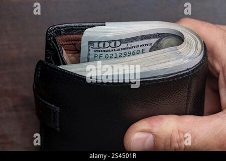 Die Hand eines Mannes hält eine schwarze Ledertasche mit einem Haufen 100-Dollar-Scheine, Banknoten, Bargeld, Nahaufnahme. Konzept für verlorenes oder gefundenes Wallet mit CA Stockfoto