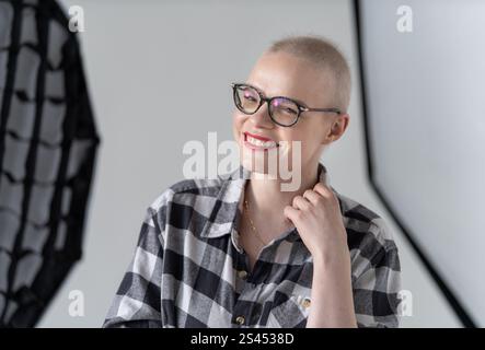 Fröhliche Person mit Brille und kariertem Hemd, die selbstbewusst in einem Fotostudio posiert. Weibliche Krebsüberlebende. Lächelnde Frau mit Brille Stockfoto