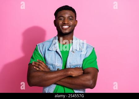 Ein charmanter junger Mann mit einer stylischen Jeansweste steht vor einem leuchtend rosa Hintergrund und strahlt Glück und Selbstvertrauen mit einem breiten Lächeln aus Stockfoto