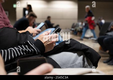 Unerkennbare Frau, die ihr Smartphone in einem öffentlichen Wartezimmer im Flughafenterminal benutzt. Nahaufnahme, verschwommene Menschen im Hintergrund Stockfoto