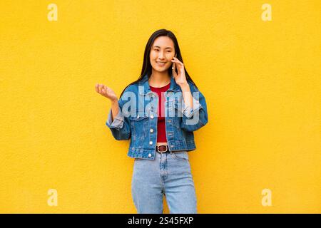 Staunte emotionale koreanische junge Frau Generation Z in Freizeitkleidung, die auf ihrem Smartphone spricht und auf einem gelben Hintergrund steht Stockfoto