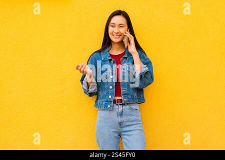Staunte emotionale koreanische junge Frau Generation Z in Freizeitkleidung, die auf ihrem Smartphone spricht und auf einem gelben Hintergrund steht Stockfoto