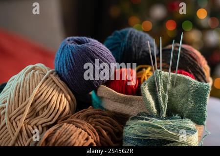 Eine Sammlung von bunten Garnbällen und Strickutensilien, die ordentlich auf einem Tisch angeordnet sind, vor einem gemütlichen Hintergrund mit festlichen Lichtern und einer Christma Stockfoto