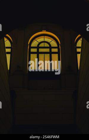 Blick von innen auf eine Treppe mit Blick auf ein hohes bogenförmiges Fenster mit warmgelbem Licht. Die symmetrische klassische Architektur und tiefe SHA Stockfoto