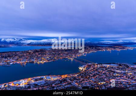 Dämmerung Luftaufnahme von Tromsø, Norwegen, im Winter. Die Lichter der Stadt funkeln unter völlig bewölktem Himmel, wobei die Brücke über den Fjord in der Ferne sichtbar ist Stockfoto