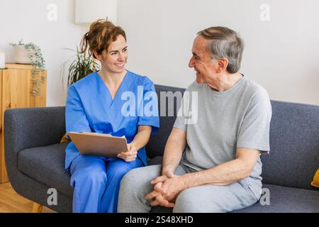 Junge Ärztin in blauer Uniform besucht einen behinderten Seniorenmann zu Hause Stockfoto