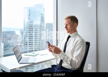 Beschäftigter junger Geschäftsmann im Büro, der am Laptop mit dem Telefon arbeitet. Stockfoto