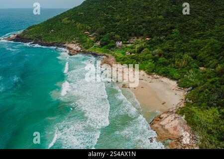 Blick aus der Vogelperspektive auf einen abgelegenen Strand mit türkisfarbenem Wasser, umgeben von üppigem Wald, felsiger Küste und verstreuten Häusern in Brasilien. Stockfoto