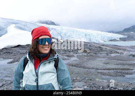 Porträt einer jungen Frau, die zur Seite schaut, eine rote Mütze, eine blaue Jacke und einen Rucksack trägt, stehend mit dem Austdalsbreen-Gletscher im Hintergrund in N Stockfoto