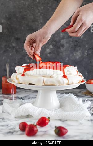 Nahaufnahme der Hände, die frische Erdbeerscheiben auf einen pavlova mit Schlagsahne und Erdbeersauce auf eine Marmoroberfläche legen. Stockfoto