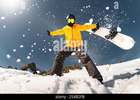 Der fröhliche aktive Snowboarder steht mit offenen Armen und hält das Snowboard in den Händen gegen den flauschigen Schnee, der in den Himmel fliegt Stockfoto