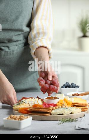 Frau serviert Antipasti-Platte mit verschiedenen Käsesorten und anderen Snacks mit Trauben an einem grauen texturierten Tisch drinnen, Nahaufnahme Stockfoto