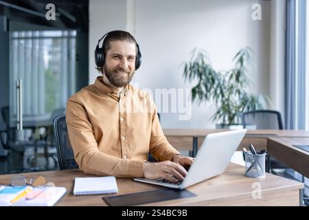 Lächelnde professionelle Kopfhörer bedienen ein Notebook an einem hellen, modernen Arbeitsplatz. Stockfoto