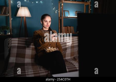Frau aus den 30er Jahren isst Popcorn und schaut Filme im Fernsehen, während sie mit der Fernbedienung die Kanäle wechselt und abends allein auf der Couch sitzt Stockfoto