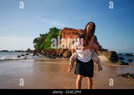 Die freudige Frau bekommt einen Huckepack vom Mann am Strand. Ein Paar genießt tropische Zuflucht, Sandstrand in der Nähe des Meeres. Spielerische Partner Punkte auf Distanz. Romantisch Stockfoto