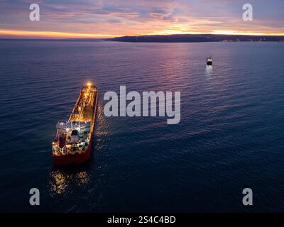 Aus der Vogelperspektive eines hell beleuchteten Tankschiffes auf dem Meer während der Dämmerung, umgeben von offenem Wasser und einem leuchtenden Horizont Stockfoto