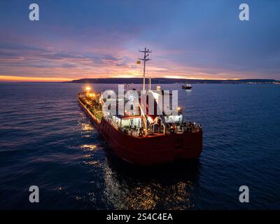 Aus der Vogelperspektive eines hell beleuchteten Tankschiffes auf dem Meer während der Dämmerung, umgeben von offenem Wasser und einem leuchtenden Horizont Stockfoto