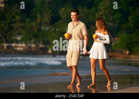 Romantisches Paar spazieren barfuß am Sandstrand, Mann und Frau in Sommerkleidung halten Hände und genießen den Sonnenuntergang. Zu einem Spaziergang über das Meer mit tropischen Getränken kombinieren Stockfoto