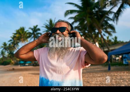Ein älterer ethnischer Mann mit grauem Bart genießt Musik am sonnigen tropischen Strand mit Sonnenbrille und Kopfhörern. Stilvolle ältere Tänze, aufmunternde Stimmung Stockfoto