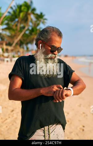 Der stilvolle ältere graue Bärenmann mit Sonnenbrille benutzt eine intelligente Uhr am tropischen Strand. Moderner aktiver Lebensstil. Älterer Mann überprüft Fitness-Tracker auf Stockfoto