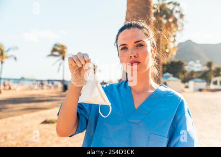 Eine Ärztin in einem blauen Peeling-Anzug, die eine Gesichtsmaske hält, ernsthafter Gesichtsausdruck Stockfoto