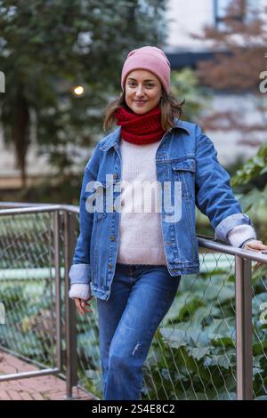 Stilvolle Frau posiert in Winterkleidung, lächelt selbstbewusst in einem Stadtpark, umgeben von Bäumen und genießt die kalte Jahreszeit im Freien Stockfoto